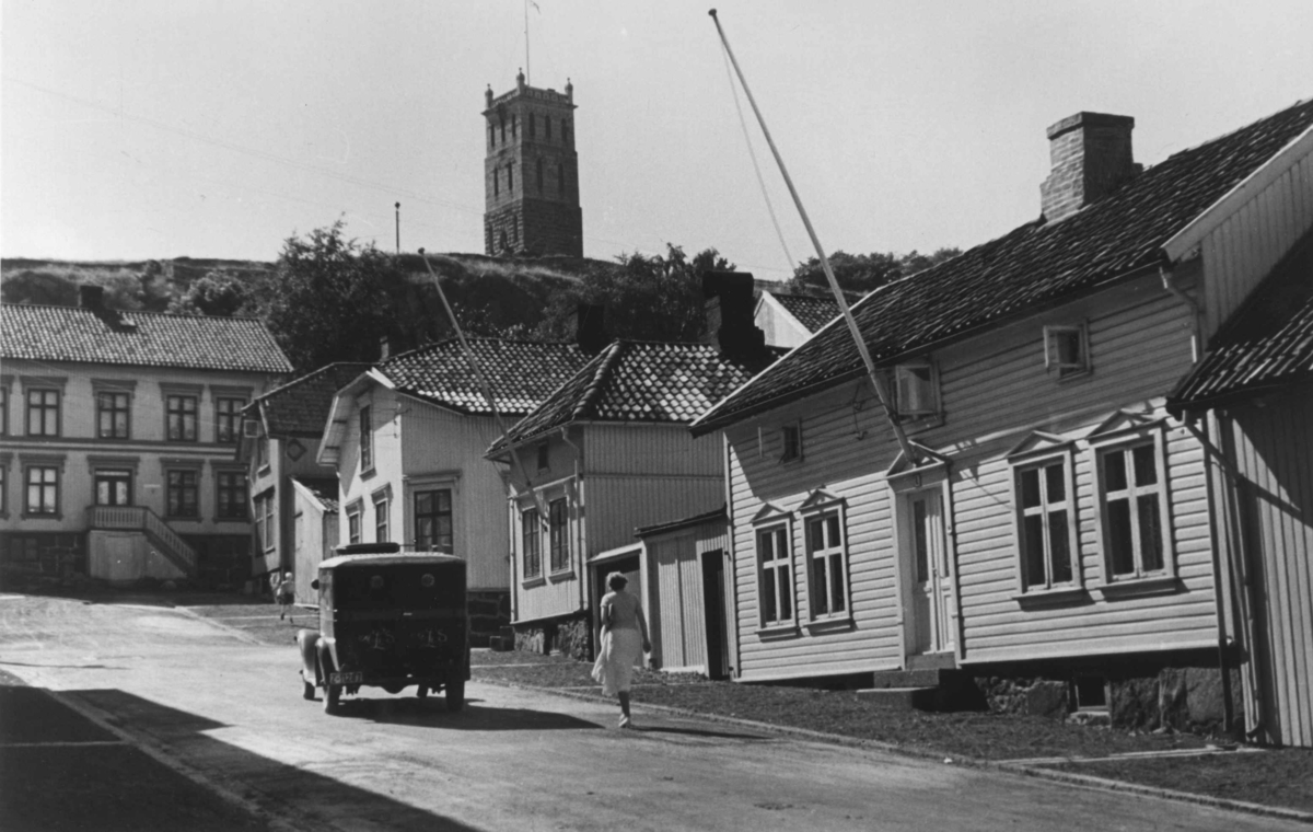 Gateliv i Tønsberg. En kvinne går og en bil kjører opp en gate blant trehusene. I bakgrunnen sees Slottfjelltårnet.
