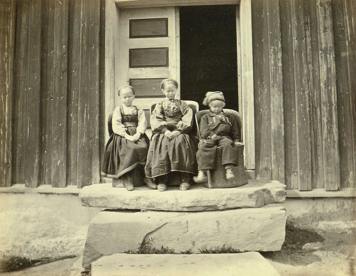 Barnegruppe i folkedrakter, Heddal, Notodden, Telemark. To piker og en gutt sitter på kubbestoler foran inngangsdør.
Fra serie norske landskapsfotografier tatt av den engelske fotografen Henry Rosling (1828-1911).