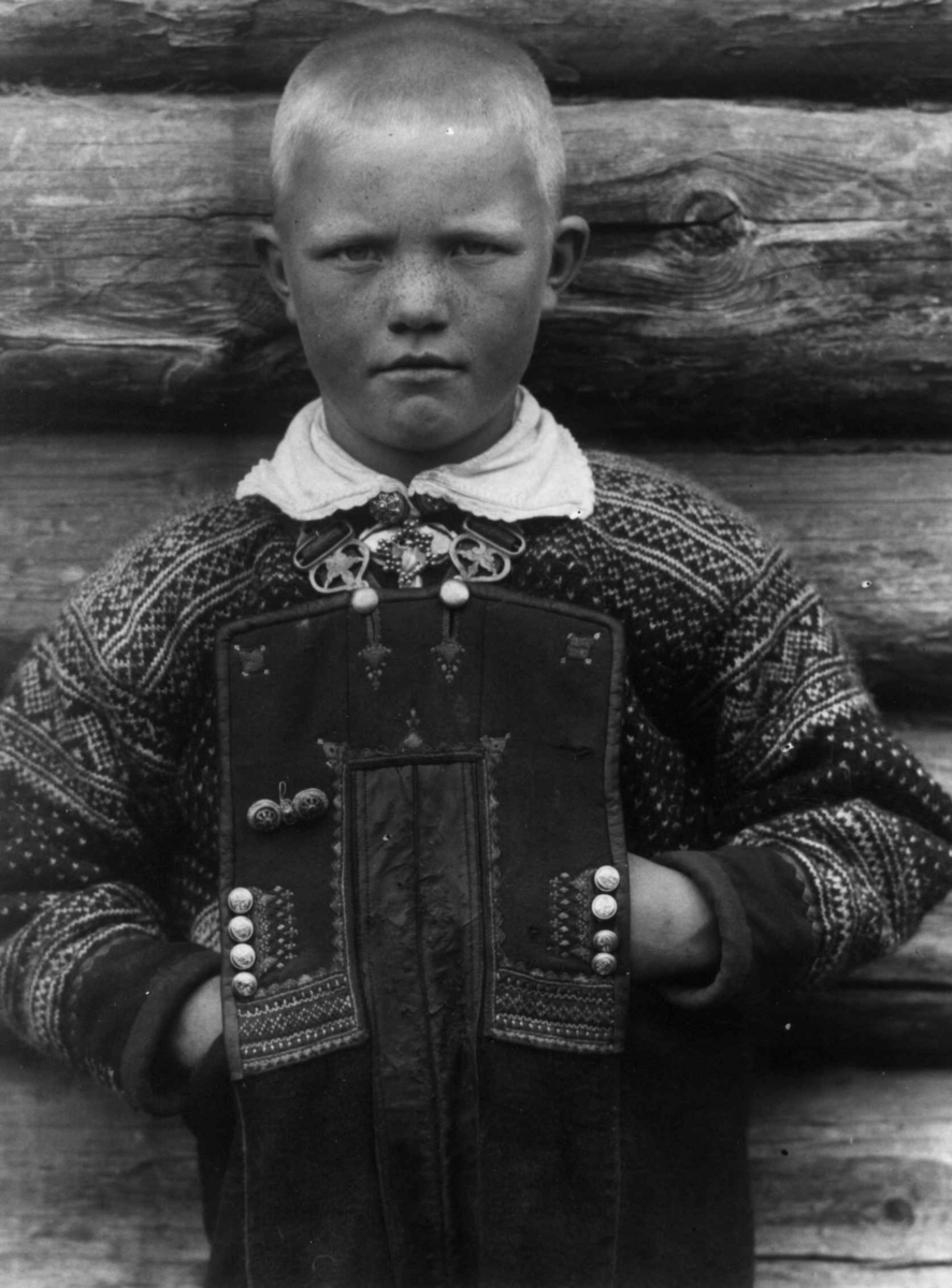 Barnedrakt, portrett, Valle, Setesdal, Aust-Agder, antatt 1924. Gutt poserer foran tømmervegg. Fra "De Schreinerske samlinger" (skal oppgis).