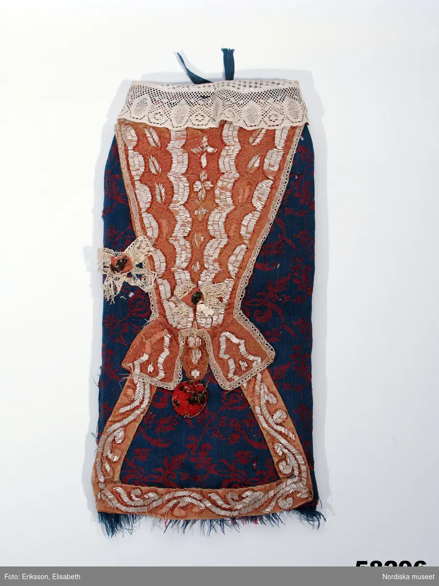 Doppåse av fint mönstervävt kamgarnsylle, kallat florett, med mörkblå botten i satin och mönsterbildande inslag i kypert i rött, i ryggen skarvat av flera bitar, sannolikt återbruk av äldre plagg, mitt fram finns ett igensytt sprund. Framsidan dekorerad med en bröstlapp från en kvinnodräkt från 1600-talet, av ljust brunorange atlassiden med ett tätt broderat gallermönster i tvinnat silke samt slingor av tätt sittande vita stavpärlor nedsydda med oblekt lingarn. Runt bröstlappen en smal knypplad uddspets. På var sida om bröstlappen och mitt på applikation av spetsrosetter med  bitar av metallspets i mitten.( Spetsarna av 1700-talstyp). Nedanför bröstlappen en rundel av rött tyg med rester av metalltråd och lintråd. Den är fastsatt med en stor ålderdomlig knappnål. Kring halsöppningen krås av vit maskinvävd bomullsspets. I nederkanten upprispad frans av florettetyget. Ofodrad.
Anm. Bröstlappen har från början varit rosaröd, men är starkt blekt. Pärlor har lossnat på flera ställen. Spetsrosett på höger sida saknas.
Hål efter skadeinsekter.
Enligt uppgift första gången använd 1730.
 Halskråset är från 1800-talet. Florette var ett fint köpetyg , sannolikt importerat. Vävdes bl.a i England. Se prov i Berchs samling. När det använts i allmogedräkter har det varit som bröllopskläder hos burgna ägare.
/Berit Eldvik dec 2005

En synål eller knappnål kunde gömmas i dopdräkten som skydd mot de onda makter som hotade det lilla hedniska barnet under färden till kyrkan, innan det genom dopet fått  kyrkans skydd.
Lena Kättström Höök juni 2008