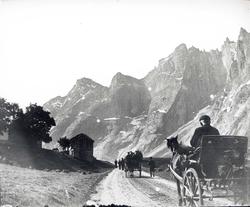 Vei med hestekjerrer mellom fjell, gård på den ene siden av 