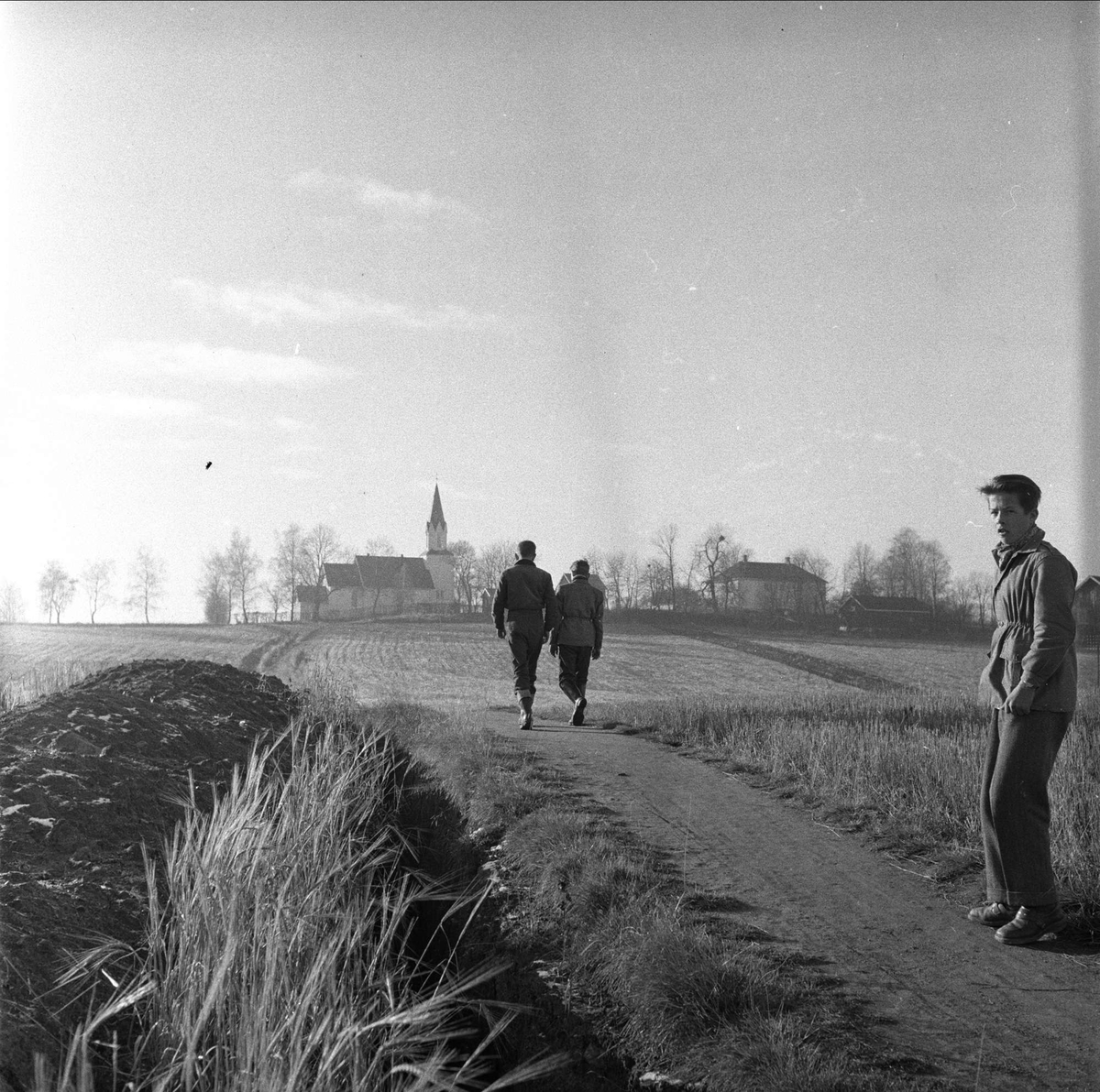 Kråkstad kirke, Ski, Akershus, 03.11.1956. Tre menn på en vei, kirken i bakgrunnen.