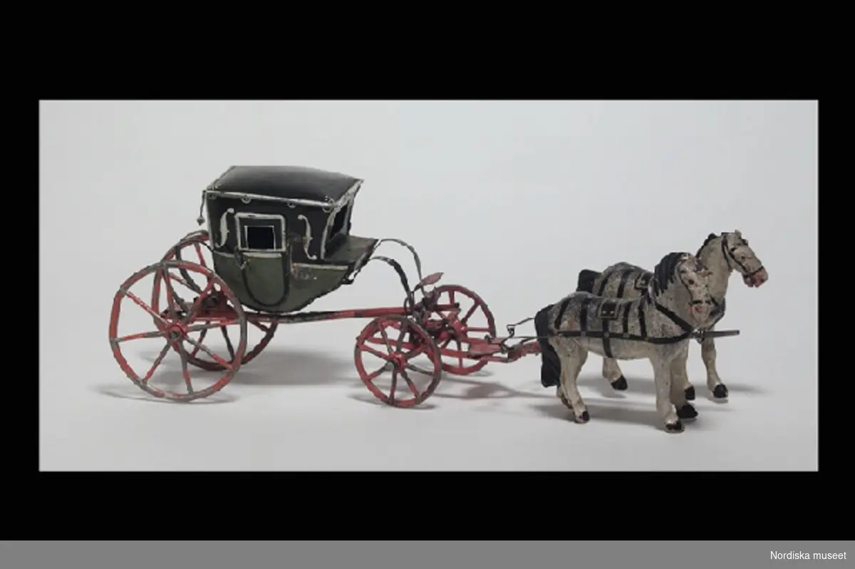 Huvudliggaren:
"a-b Leksaksvagn, kupévagn på c-fjädrar m. 4 hjul, av plåt, grön o svartmål. m. röda hjul, samt parhästar av trä, defekta. G[åva] 28/2 1927 [av] Fröken Elisabeth von Feilitzen, Stockholm."

Inventering Sesam 1996-1999:
L 27    B 6,5    H 9  (cm)
a) vagn  b) hästar
Ekipage, leksak, kup¿vagn, sk berlinare, förspänd av två hästar. Sluten vagnkorg av järnbleck, målad i svart och grönt, framtill med plats för en kusk, två öppningsbara dörrar. Underrede av järnbleck, målat orange, hjul med ekrar, vridbart främre hjulpar, tistelstång. Två hästar skurna i trä, målade i vitt med svarta manar, fastsatt med tistelstången medelst stag av järn.
Vagnkorgen undertill märkt: "M" ovanför "CB", eventuellt = Büchner.
Anm: Ena hästen med avslaget bakben.
Jmf snarlika hästar till ekipage  inv 71.065.
Birgitta Martinius mars 1998