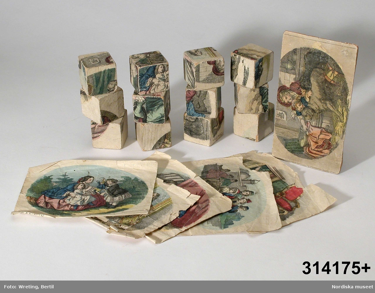 Katalogkort:
"Läggspel bestående av 12 st kubiska klossar med en klistrad del av färgbild på varje sida. Dessutom 5 st mtsvarande färgbilder samt en dito fastklistrad på en träplatta (locket till den ursprungliga asken.)
