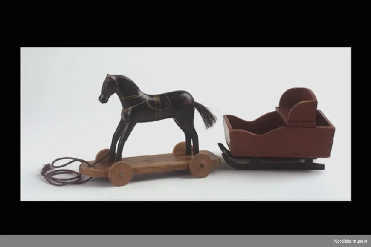 Inventering Sesam 1996-1999:
L 27,5, B 10, H 21 cm (häst)
L 21, B 14,5 cm (släde)
Leksaksekipage, häst och släde. Handgjort av trä och målat. Svartmålad häst med gulddekor, tagelsvans, stående på en träplatta med fyra hjul. Dragsnöre i fram. Brunmålad släde med svarta medar.
Tillverkad i trä av givarens morfar, verkmästare vid Gävle Dala Järnvägar, August Fredrik Hoffman, Södra Hospitalgatan 10, Gävle, f. 1857. Han var en händig hantverkare enligt givaren.
Julklapp till sonen Eskil Fredrik Hoffman, f. 1890 d. 1903. Till jul fick syskonen var sin present - endast en och ej paketerad. Aldrig gåva till födelsedag eller namnsdag.
Givaren lekte själv med ekipaget -  ute under vintertid - vid besök hos mormor i Gävle. Kopplade ihop med ett snöre i slädmedarna och en ögla om hästens hals.
Medarna är utbytta någon gång under 1970-talet.
I R B 1993 [=Ingrid Roos Björklund]