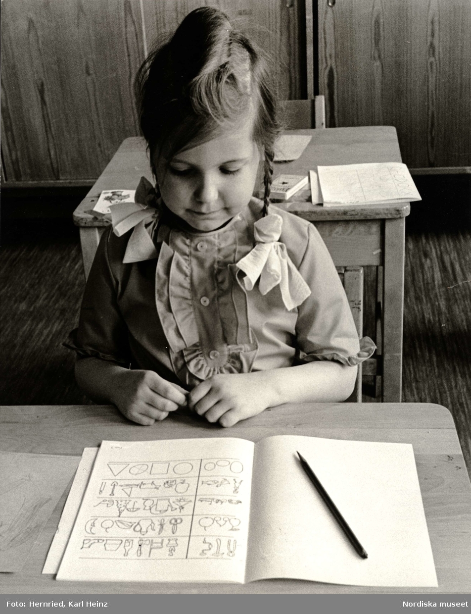 Skolmognadsprov. Flicka i skolbänk av trä med övningsbok och penna framför sig