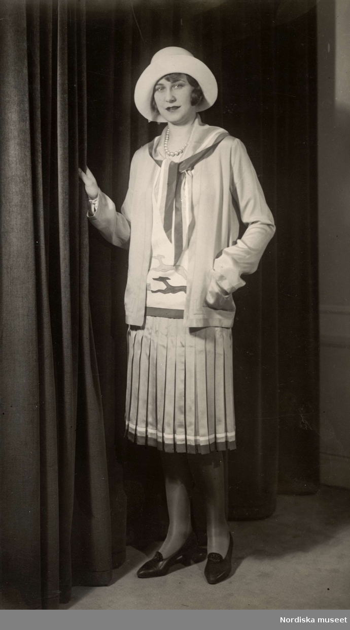 Modevisning i mars 1929. Kvinna iklädd dräkt med plisserad kjol och hatt. Nordiska Kompaniet.