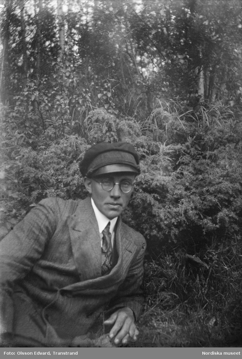 Edvard Olsson i kostym, vegamössa och glasögon ute i skog och mark. Självporträtt.