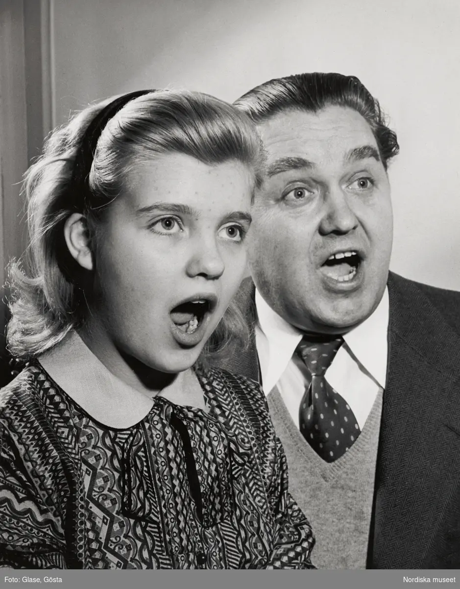 Porträtt av den svenske operasångaren, tenoren Jussi Björling (1911-1960) och hans dotter Ann-Charlotte (f. 1943), även hon sångerska, framförallt inom operettens värld. Far och dotter ses i bröstbild, de sjunger tillsammans. Ann-Charlotte bär en mönstrad blus eller klänning med krage, Jussi Björling är klädd i fiskbensmönstrad tweedkavaj, slipover, skjorta och prickig slips.