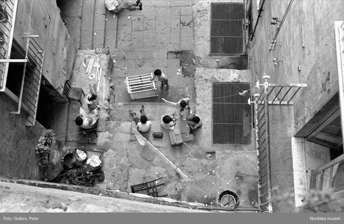 Fågelperspektiv. Bilden tagen rakt uppifrån ner på gata eller gård. Barn som leker. Kvinna som sitter på stol. Italien