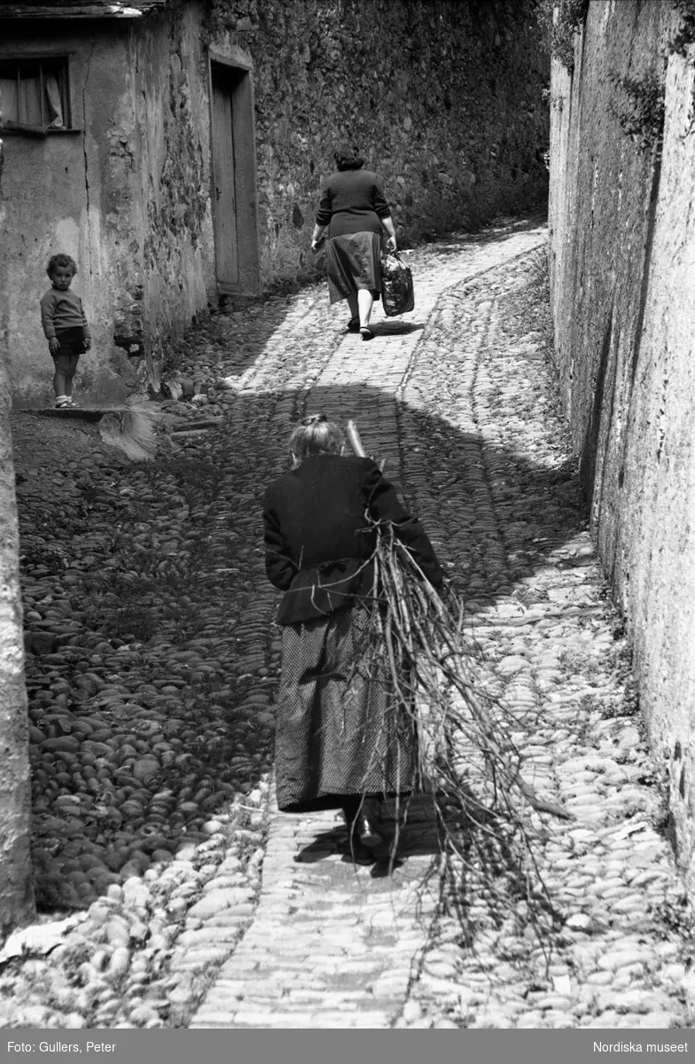 Kullerstensgränd. Två kvinnor går uppför en backe med ryggen mot kameran. Den bortre kvinnan bär en kasse i höger hand och har just passerat en liten pojke som tittar ner mot kameran. I förgrunden ser man den äldre kvinnan som släpar på en fång grenar och kvistar. Levanto, Italien