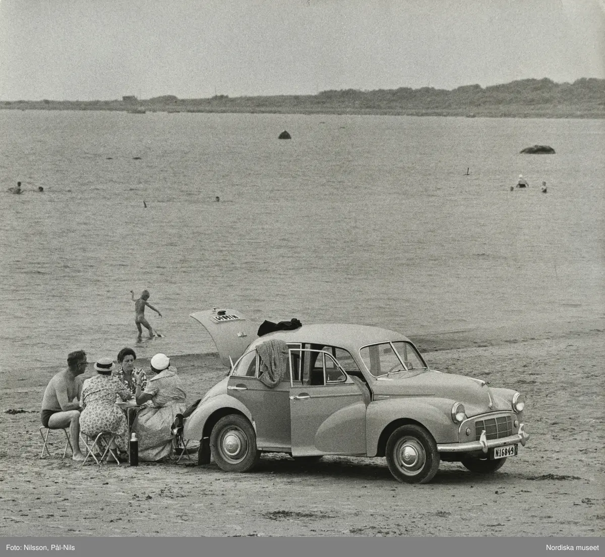 En bil, Morris Minor, parkerad på badstrand vid Frösakull, Halland. Tre kvinnor och en man sitter vid picknickbord, badande pojke i bakgrunden.