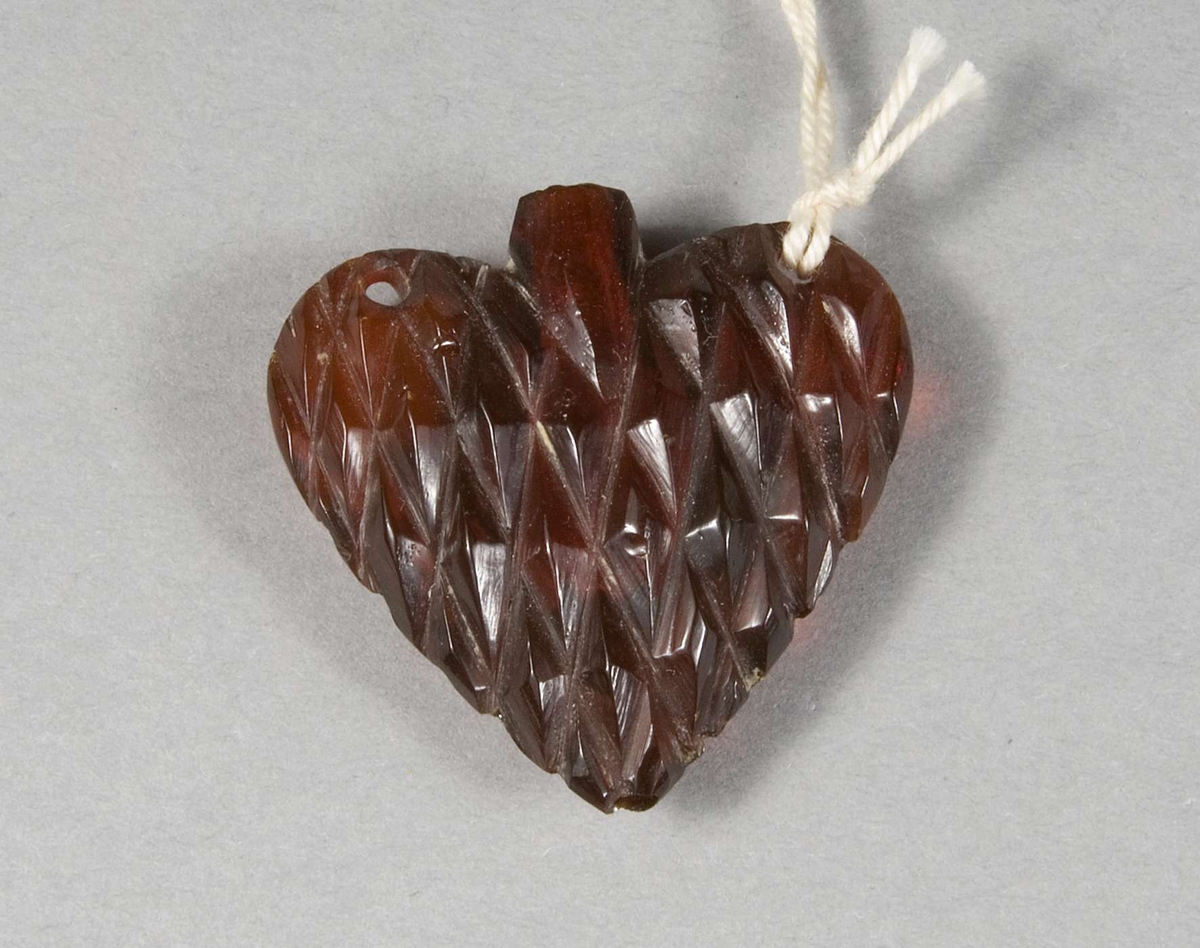 Hjärtformat smycke av mörk bärnsten med inskription: Glöm icke givaren. Två hål för upphängning. Hjärtat har en flaskhalsliknande öppning som leder till en hålighet.