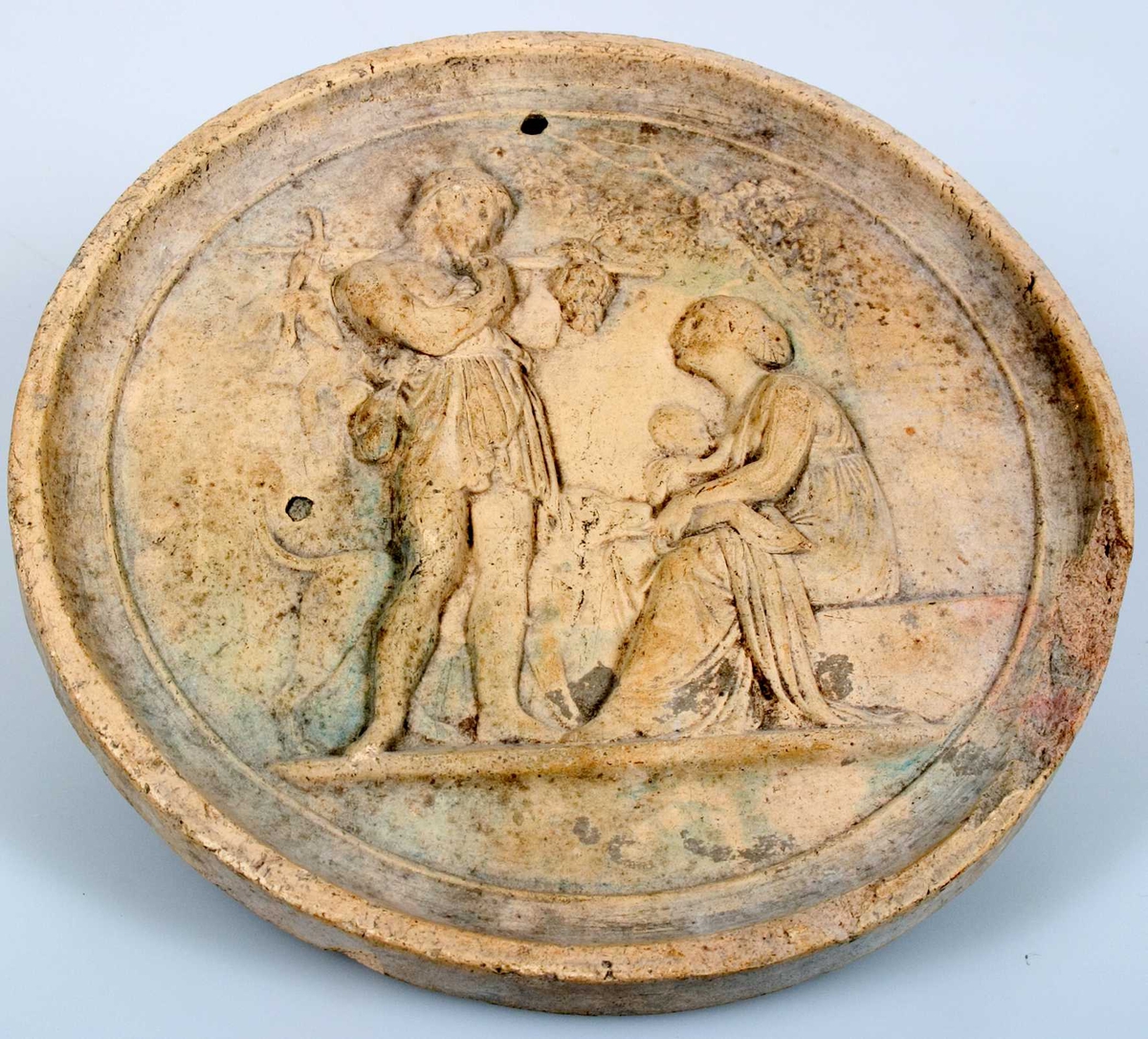 Medaljong, rund av gulaktigt lergods med klassisk figurscen i relief. På baksidan stämplad CAB.