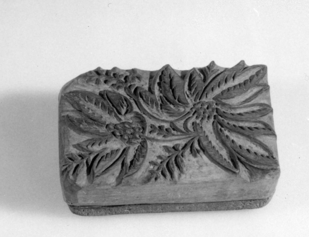 Tryckstock av två sammanfogade trästycken, för tapettryck.
Skuret motiv i form av fyra stiliserade blommor med slingor emellan.
