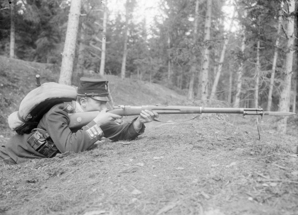 En armésoldat lägger an ett gevär liggandes på en kulle. Mannen är Ernst Axfors och tillhörde I 4 regemente.