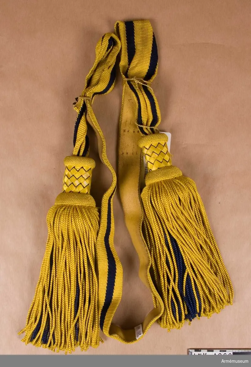 Paradskärp tillverkat i språngningsteknik. I enkel bredd utgörs randningen av två mörkt blå och tre gula ränder. När skärpet vikes dubbelt, som det görs vid användning, är en blå och två gula ränder synliga. Insidan skodd med gult läder. Två mässingspännen med vardera fyra torn. Tofsens frans består av gula och blå silkesnoddar med det blå längst in. Huvudet är klätt med gult silke vilket är spunnet över en pappremsa och sedan flätat över tofshuvudet.