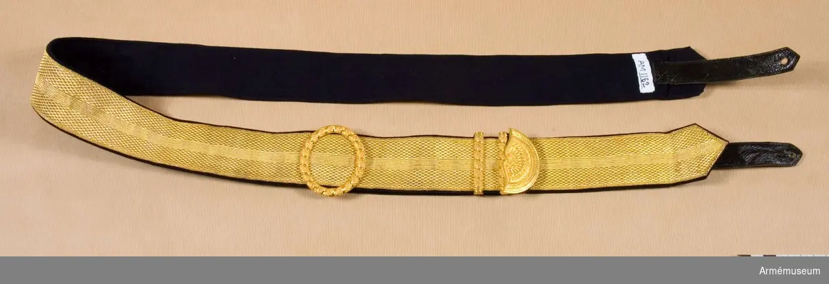 Av guldgalon m/1895, 45 mm bred, fodrad med mörkblått kläde. Ciselerade beslag för att avpassa längden. I ändarna lädertampar för fastgörning vid kartuschväskan.