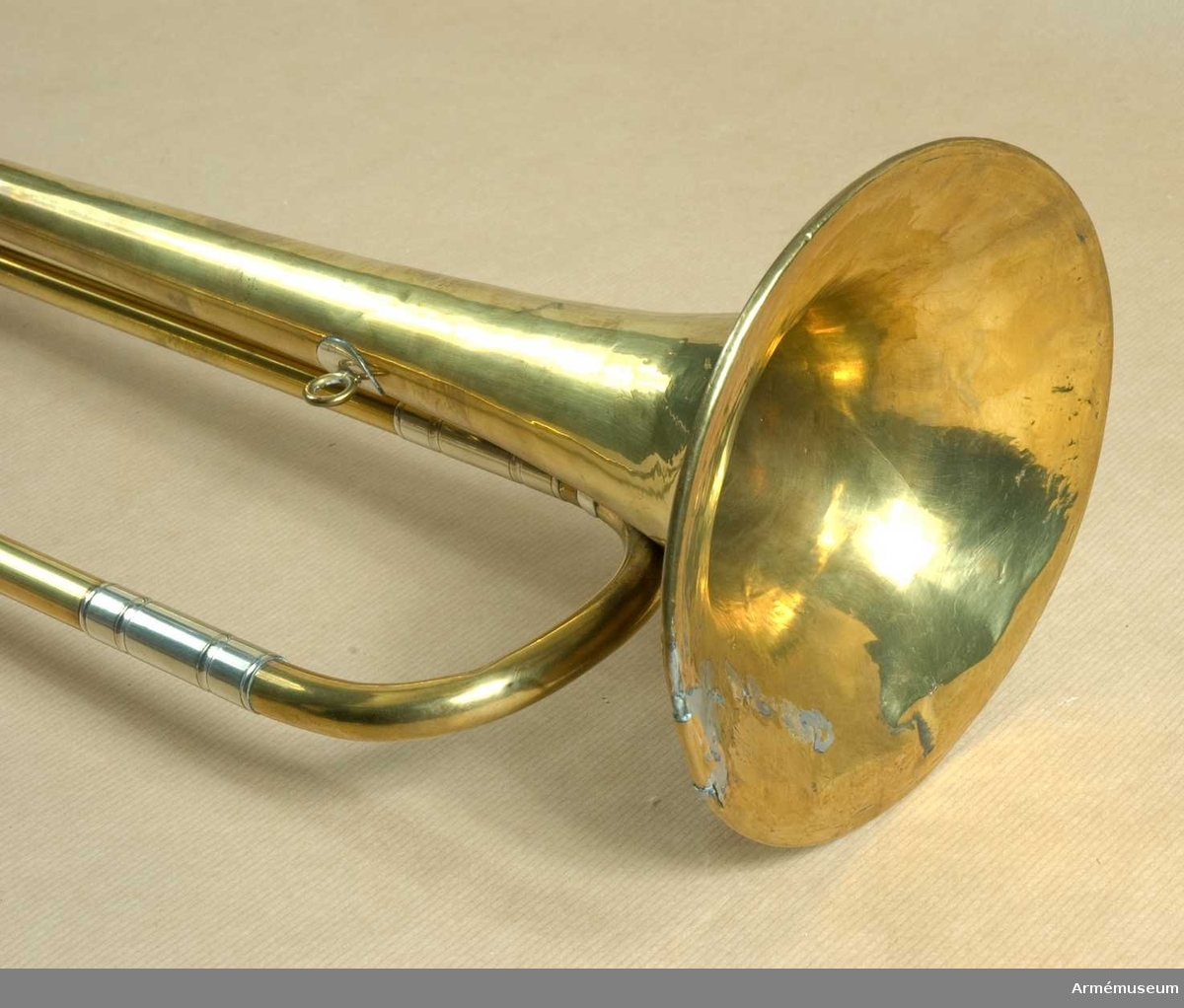 Fanfartrumpet med samhörande fana. Av mässing och vitmetall med en vindling. Trumpeten är signerad på klockstycket: Bohland & Fuchs, Graslitz. Höjd utan munstycke 69 cm, med d:o 72 cm.