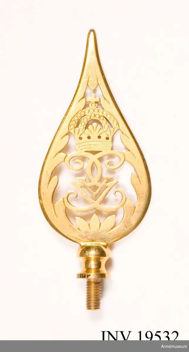 Fanspets, Gustav V:s monogram. Spetsen är tillverkad av förgylld mässing. Den har Gustav V:s spegelmonogram under en kunglig krona inramat av ett bladverk som avslutas i en smal spets. Spetsen är försedd med skruv.