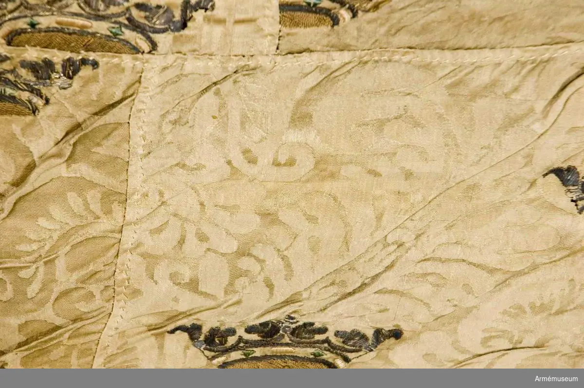 Fragment av en fana i vit sidendamast med broderade kronor. 
Duk: Tillverkad av enkel vit sidendamast. Damast från 1600-talets mitt.

Dekor: Broderad med strödda öppna guldkronor med stenar av grönt och svagt rosa silke, kronfodret gulaktigt. Duken i nedre delen fäst vid stången över linnetyg som ligger närmast stången. 

Stång: Tillverkad av furu. Nedanför dukens fäste lindat ett mönstervävt band i ca 470 mm fastsatt med tennlickor. 

Märkning på stången, inristat högst upp, strax nedanför dukens nedre kant: "K". Längre ner: "E.A.V.H" och "F.B.  ,   W". Längst ner inristat på stången sju stycken streck (IIIIIII).

Duken endast fragmentarisk. Damastens stadkant grön.