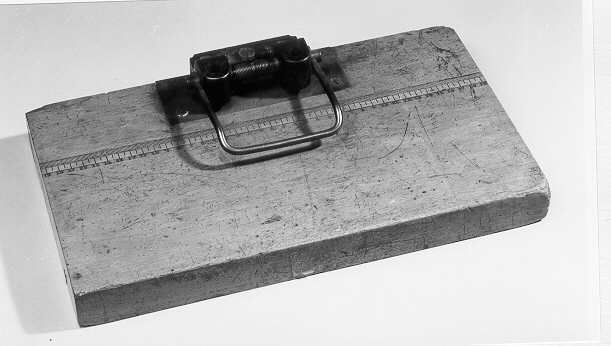Hålslagningsapparat i metall för två hål, fastskruvad i ena kanten på en träplatta. Träplattan är försedd med en centimeterskala där 0 är placerat i mitten och skalan löper 19,5 cm åt respektive håll. Funnen i förrådsutrymme på Vasagatan 8-10, 6 tr. 1982.