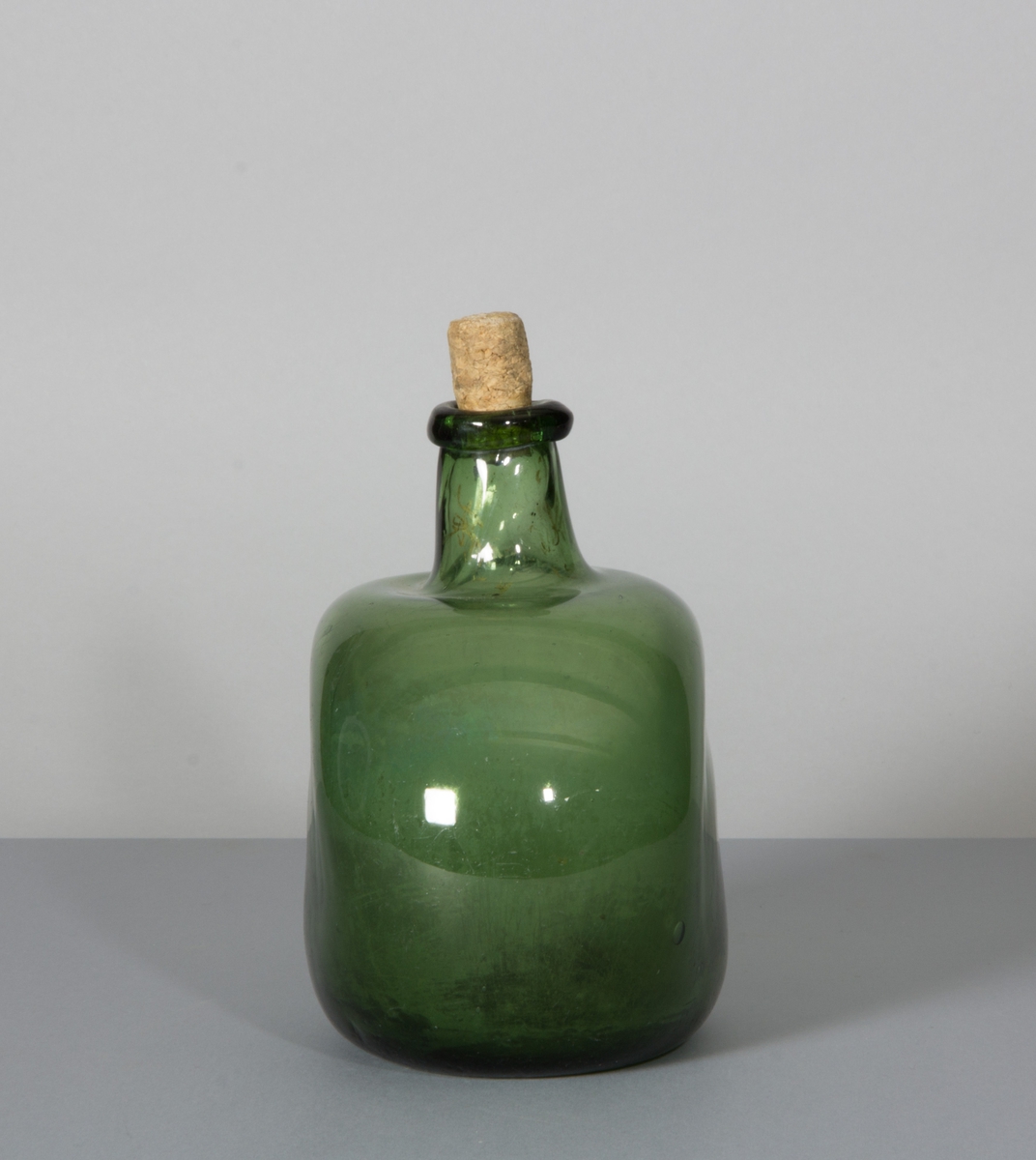 Flaska av glas, grön till färgen. Rundade sidor, hals med halsring och platt botten.