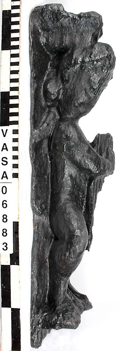 Skulptur föreställande en barnfigur spelande en harpa.
Instrumentet hålls med bägge händerna. Huvudet är vridet något åt höger. Kroppstyngden vilar på vänster ben medan höger ben är böjt. Vid höger sida syns en mantel. Ovanför hjässan sitter en snedställd äggstavslist. Figuren står på ett minimalt underlag som troligen avser att föreställa ett stycke naturlig mark. Skulpturens baksida är slät.
Skulpturen är mycket sliten.

Text in English: This is a sculpture of a child playing a harp.
The instrument is held in both hands. The head is turned slightly to the right. The weight of the body rests on the left leg, the right leg is bent. A mantle is to be seen to the figures right side. Just above the head the back board comes foreward at right angles and the front of the board is wide and is decorated with an egg moulding design which is placed diagonally across it with the right-hand side lower than the left-hand side, when the sculpture is viewed from the front. The figure stands upon a small flat plinth. The back of the sculpture is smooth.
The sculpture is very  worn.