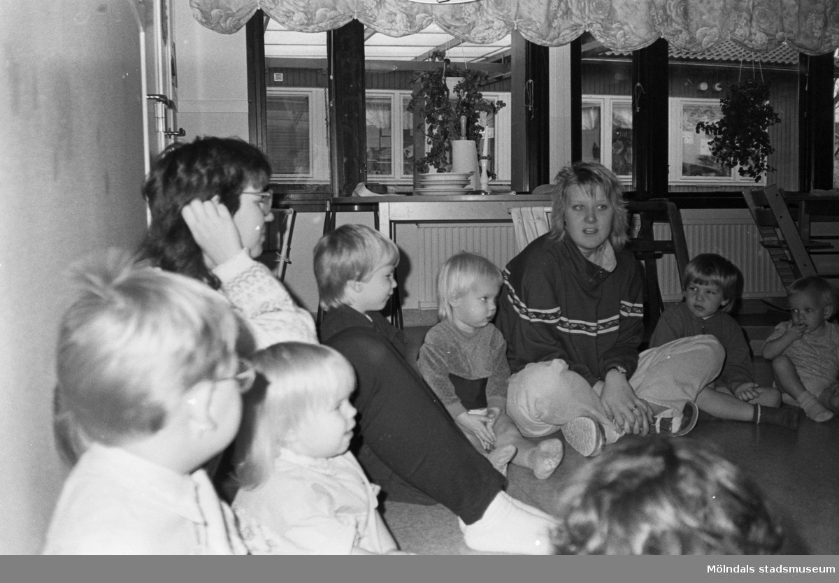 Två förskollärare och flertalet småbarn sitter tillsammans i en cirkel på golvet. Utanför rummets fönster kan man se en annan närliggande byggnad. Katrinebergs daghem, 1992.