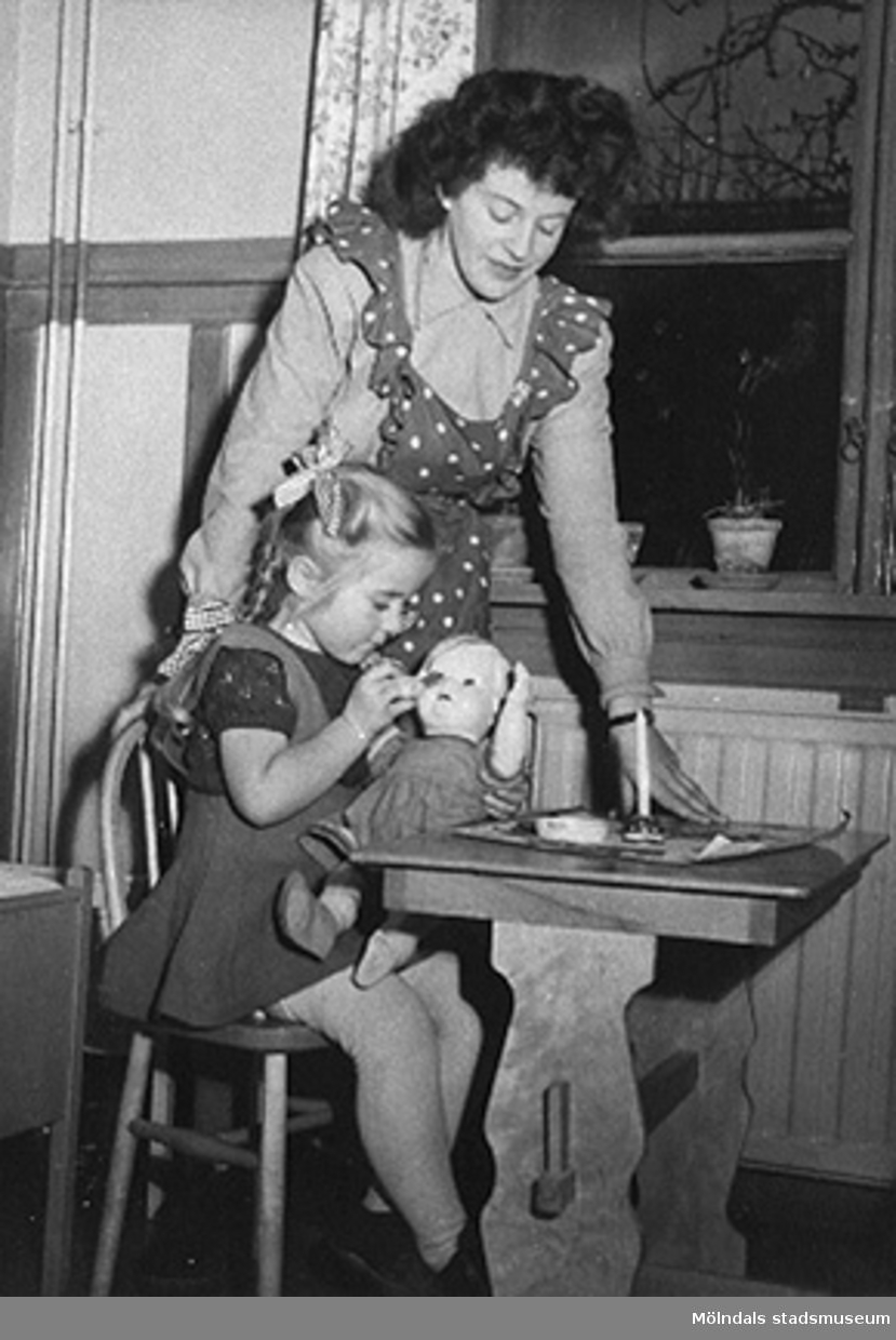 Bitr. föreståndare Margit Emilsson (gift Wannerberg -52) tittar på när en flicka pysslar om en docka, Krokslätts daghem år 1949.

Margits rödprickiga förkläde är av samma tyg som gardinerna bakom henne. Tyget är från Mölnlycke fabriker.