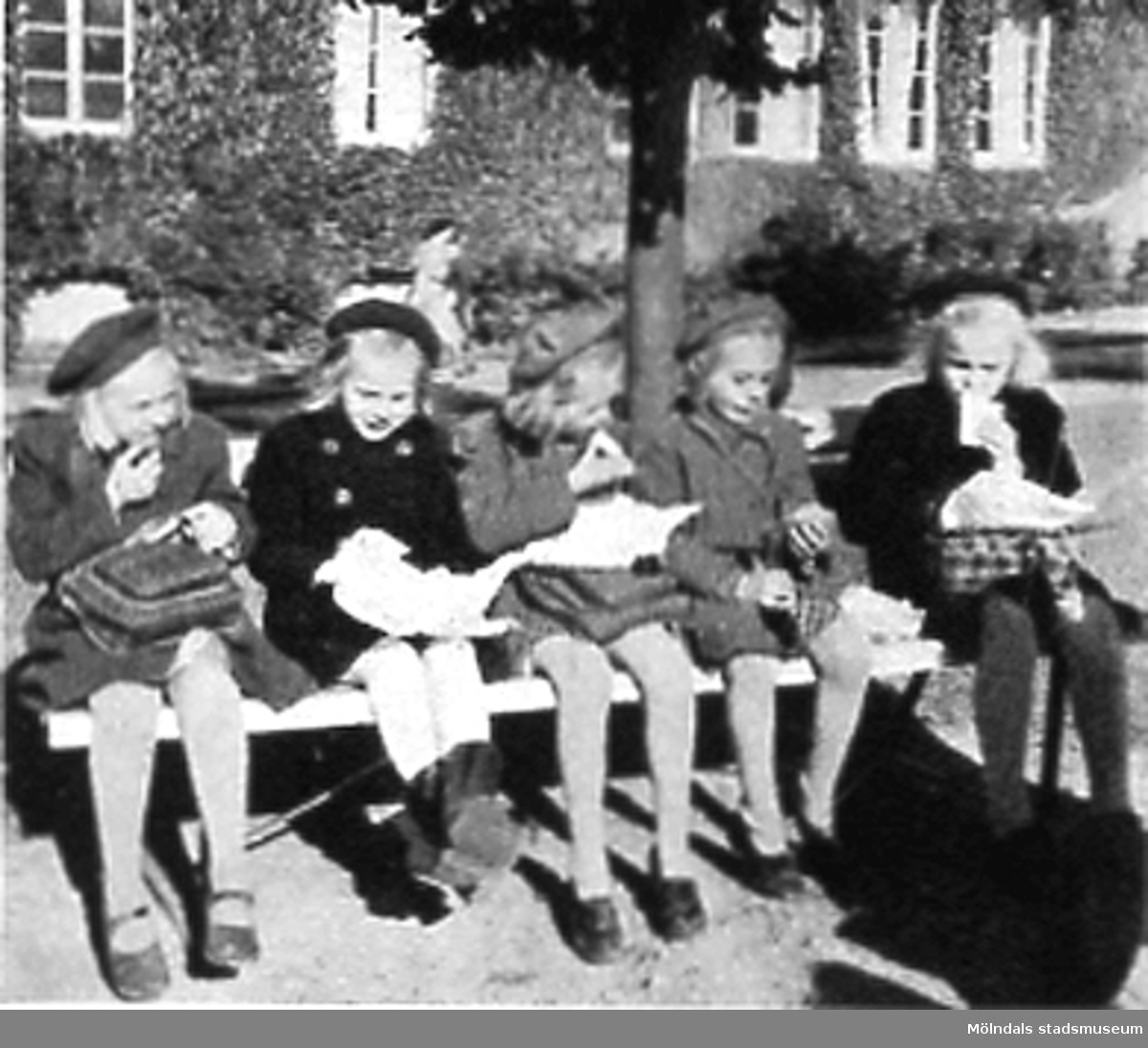 Fem flickor sitter på en bänk och har matrast utanför Kvarnbyskolan (Centralskolan), okänt årtal. Avfotograferad ur "Mölndal 1922 - 1947".