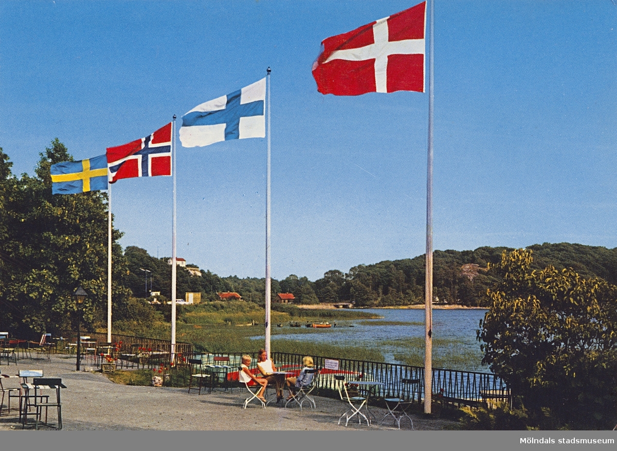 Ett vykort med utsikt över Stensjön, omkring 1967. Ett sällskap sitter och fikar vid fyra flaggstänger med flaggor från Sverige, Norge, Finland och Danmark. Bak på kortet står anteckningen: "26/6-67 1.000".