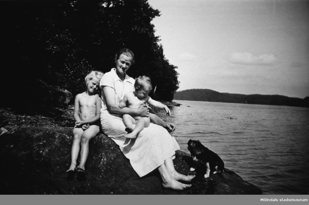 En kvinna med två barn och en liten hund sitter på en klippa, troligtvis vid Tulebosjön cirka 1930.