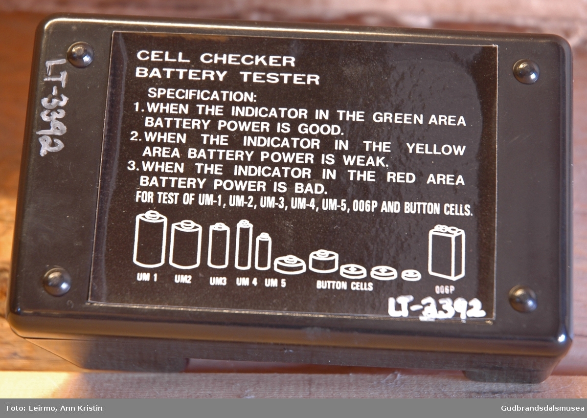Liten svart batteritester i hardplast, rektangulær, liggende. Har indikatorpanel i rødt, gult og grønt. Spor til batteriene som skal legges ned og sjekkes for strømnivå, med + og  - pol. Spesifikasjon på baksiden forteller hva indikatoren viser/betyr, viser også bilde på ulike typer batterier/celler som kan testes med dette instrumentet.