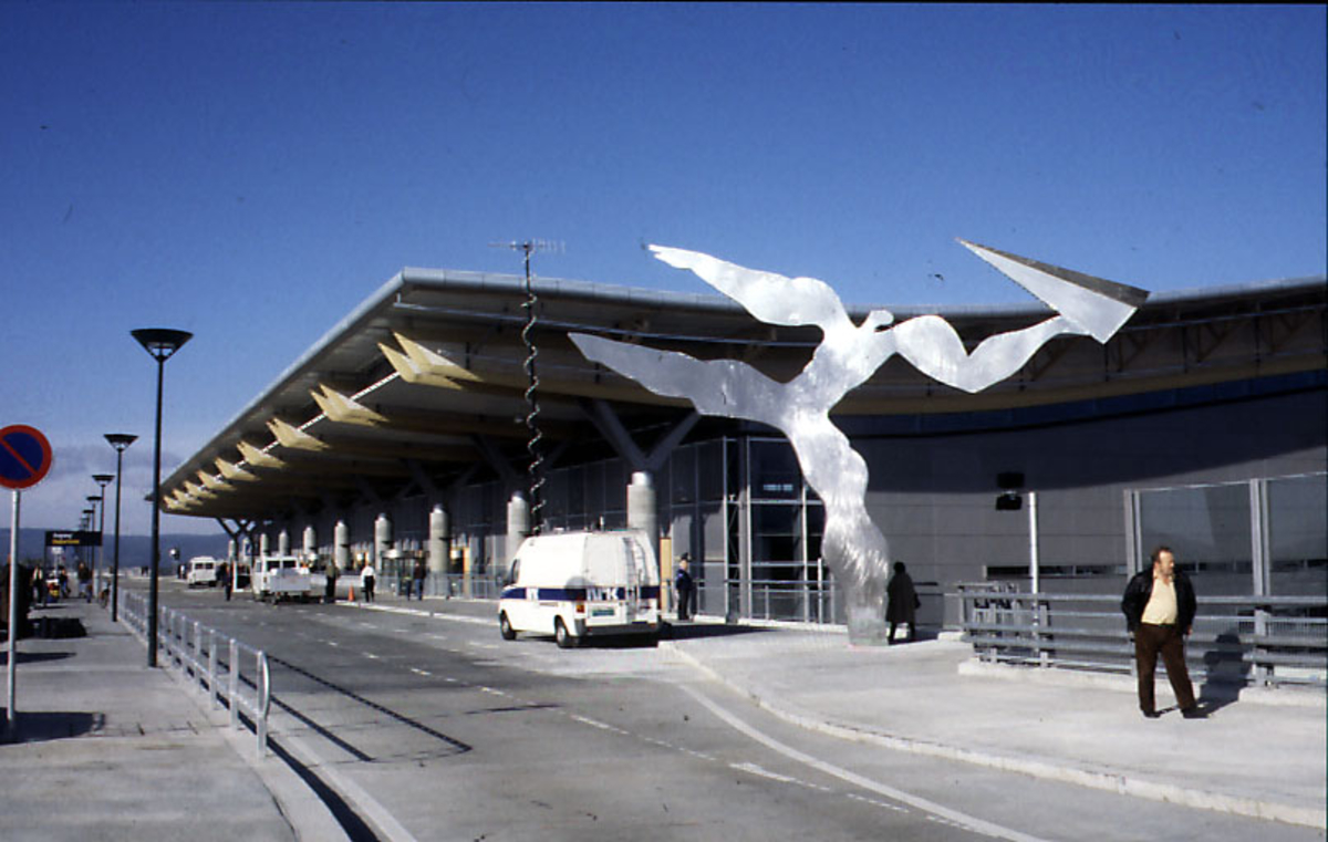 Lufthavn, flere personer, 1 skulptur og kjøretøyer ved inngangspartiet til terminalbygningen. 