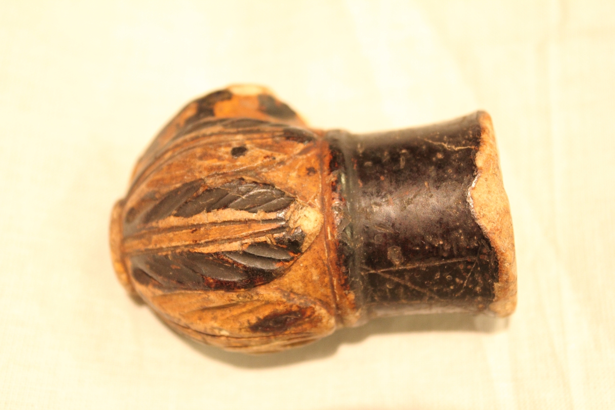 Sjølve pipa mangler. Laget av bein og har utskårne blader nederst. Øverst en rund brun ring, har ellers en brun-gul farge.