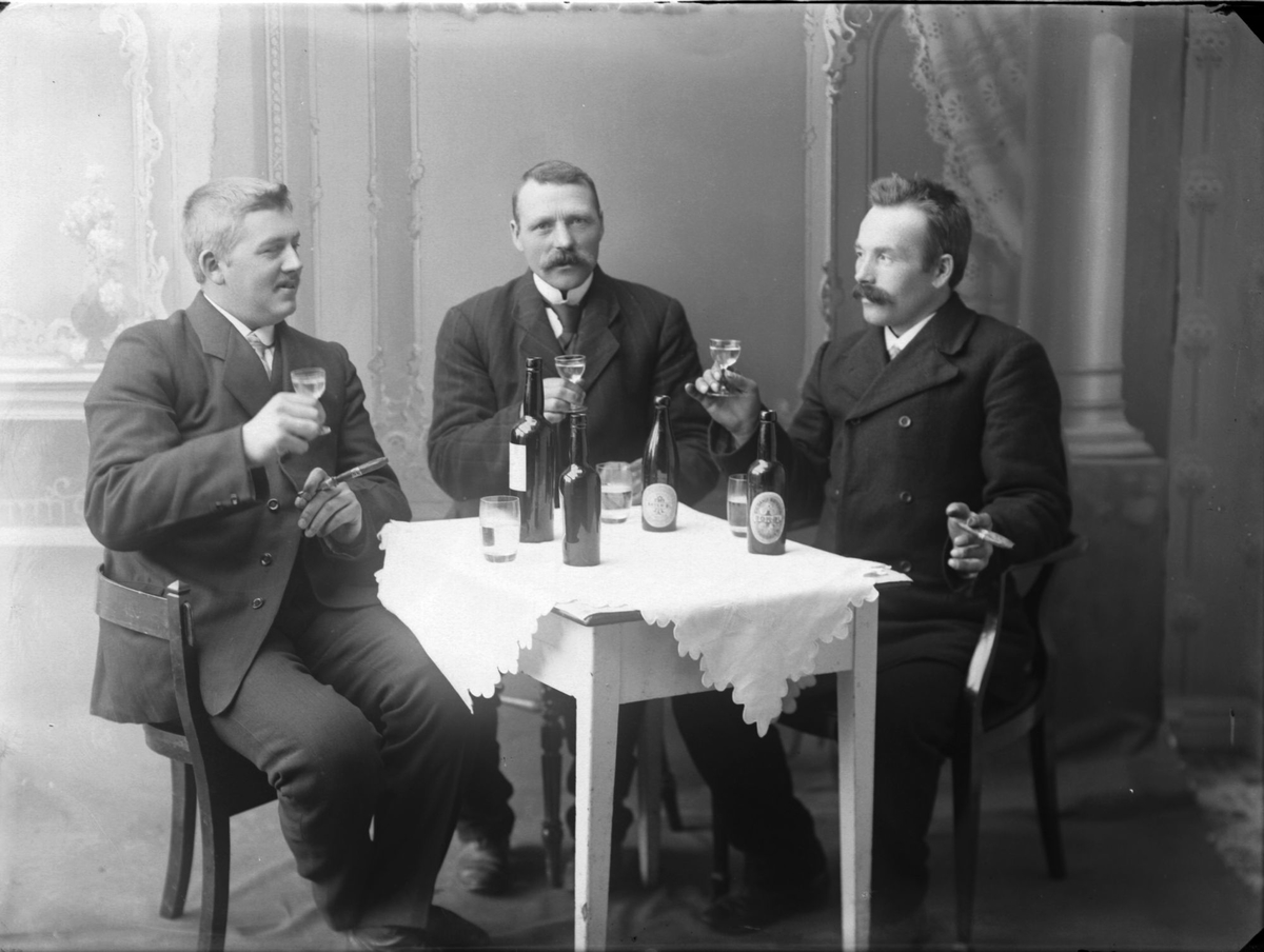 Studioportrett av en gruppe menn rundt et bord som drikker øl og vin og røyker sigar.