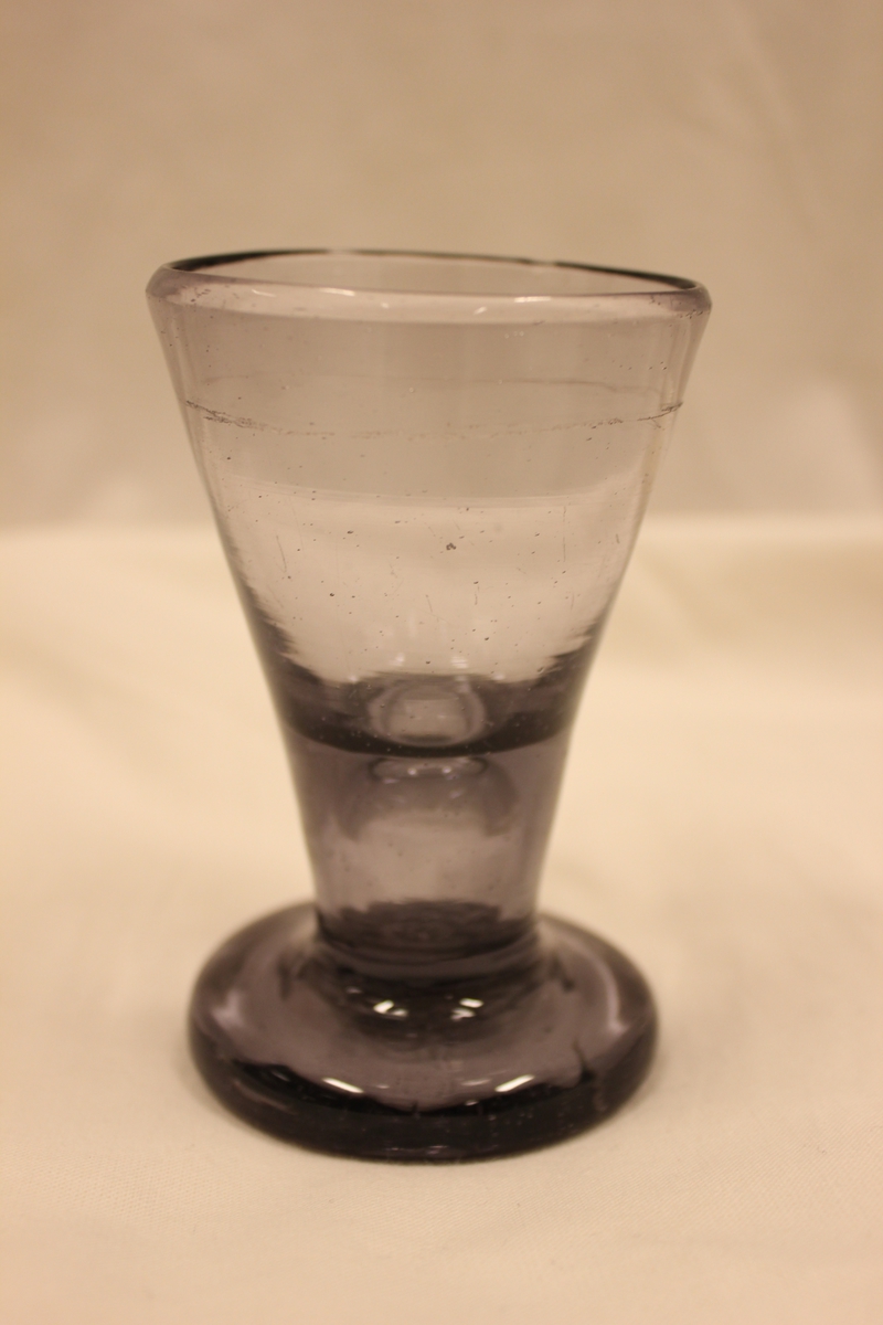 Ett drikkebeger i glass. Dette er et norsk "FRIMURER BRENNEVINS GLASS" ofte populært kalt "nøgen(naken) jomfru". Pga. fargen er det sansynlighvis fra siste halvdel av 1700- tallet. (Typen glass ble produsert langt opp i 1800- tallet). Glasset ble framstilt ved alle norske glassverk med såkalt "hvitglass" produlsjon, hvor det ble produsert klart glass i motsetning til grønt glass til flasker og vinduer. Glasset var datidens foretrukne brennevinsglass overalt i nordeuropa. De kalles "frimurerglass" i de fleste kataloger og prislister, - dette skyldes at frimurerne hadde en skikk å banke glasset hardt i bordet etter en skål, - dette krevde da en kraftig og solid fot så glasset ikke gikk i stykker. I England kalles de "firering glass" og var ofte brukt i marinen og i militæret. Når alle hadde skålet og drukket, banket de glasset hardt i bordet og det lød som en "geværsalutt!"