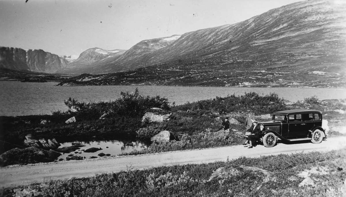 To kvinner og en mann ved bilen under biltur over Grotlifjellet, Geiranger, 1934. Sjø og fjell i bakgrunnen.