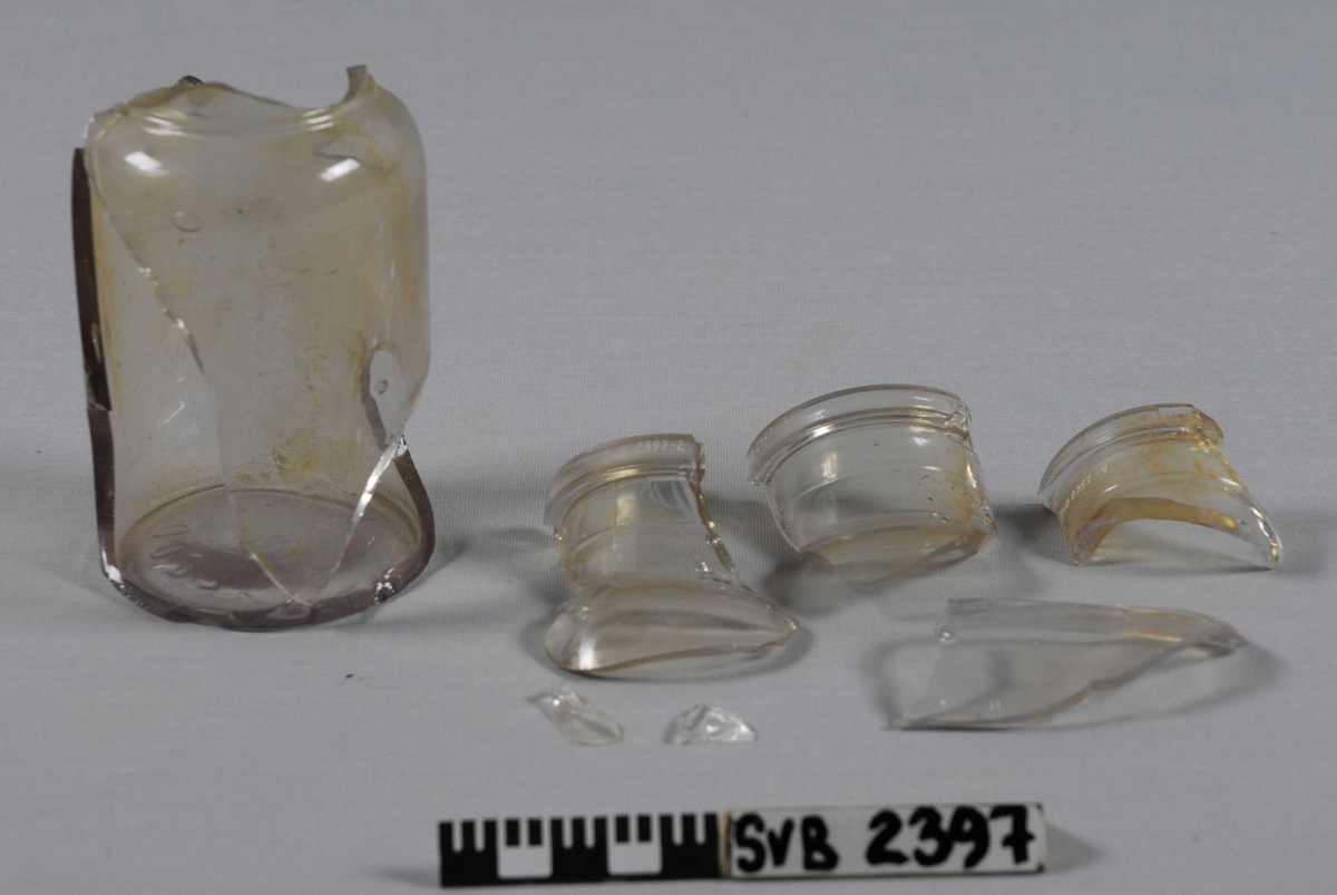 Flaske i klart glass. Tydelige luftbobler i glasset. 4 større fragmenter. En pose med mindre fragmenter. 