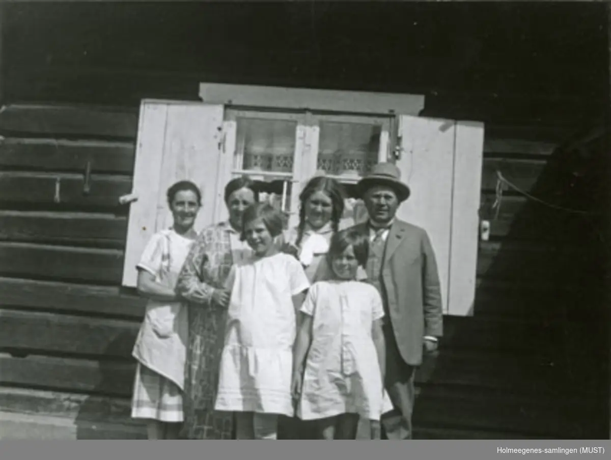 To kvinner, tre jenter og en mann står ved en husvegg. Rett bak dem et vindu med skoddene åpne. Se også ST.K.HE 2007-011-0111 til -0126.