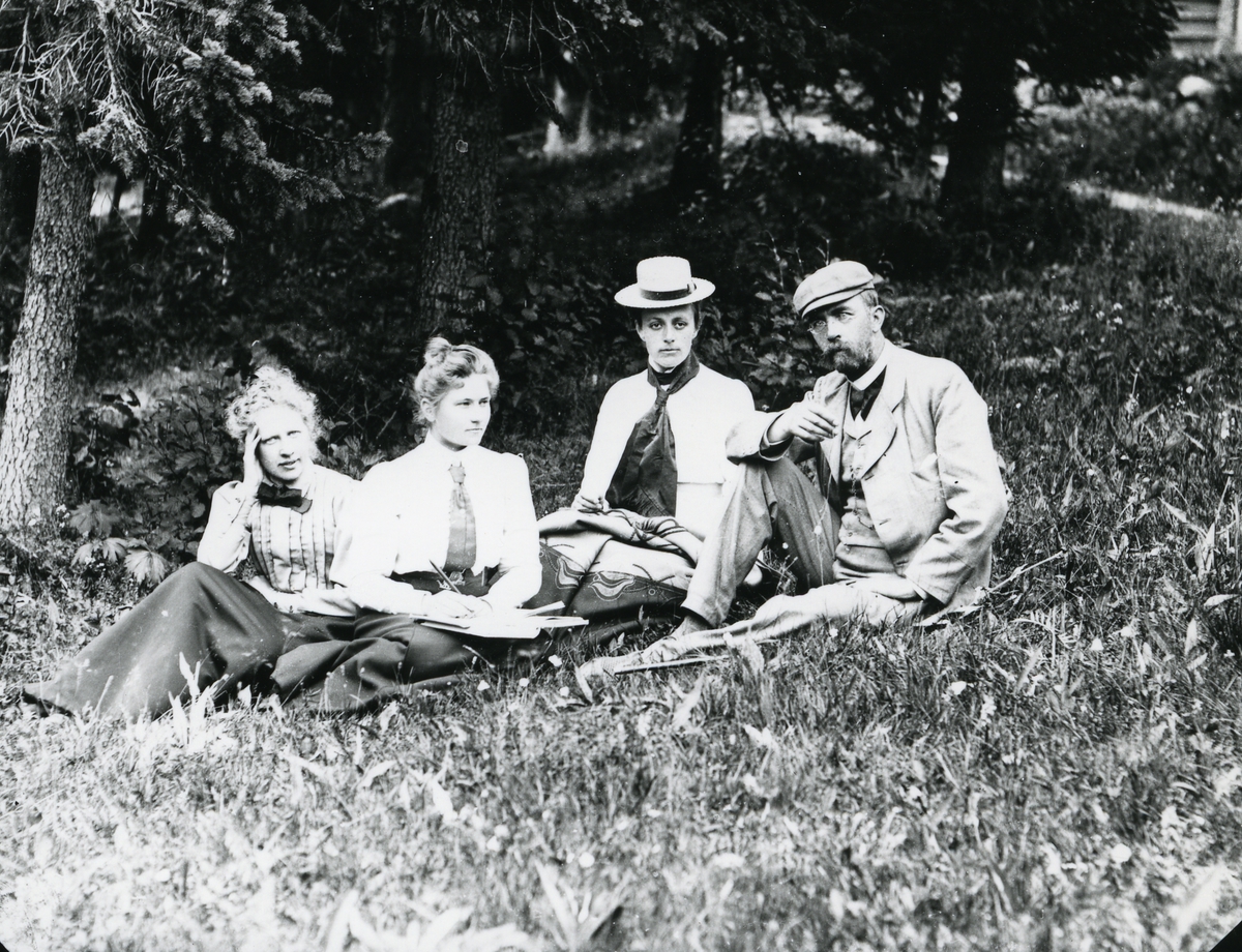 Mann og tre kvinner, sittende i gress foran skog og vei