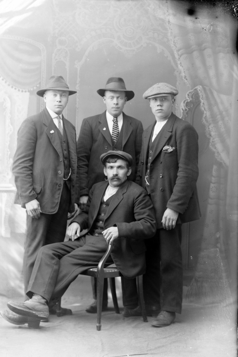 Studioportrett av tre menn stående bak én som sitter.