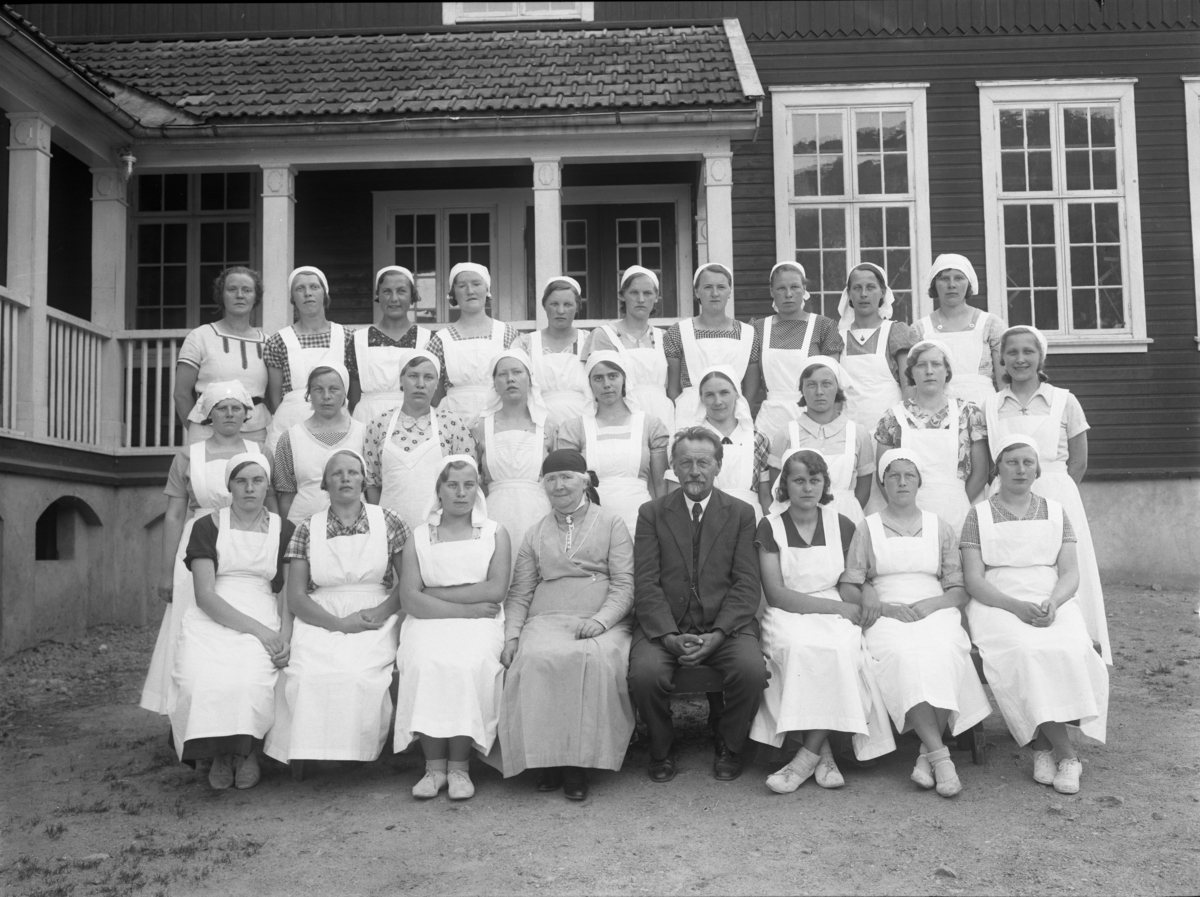 Utendørs gruppebilde av unge kvinnelige elever på husstellkurs. Folkehøgskulen/Folkehøyskolen, Utsund, Kviteseid. Jacob Naadland med frue i midten.