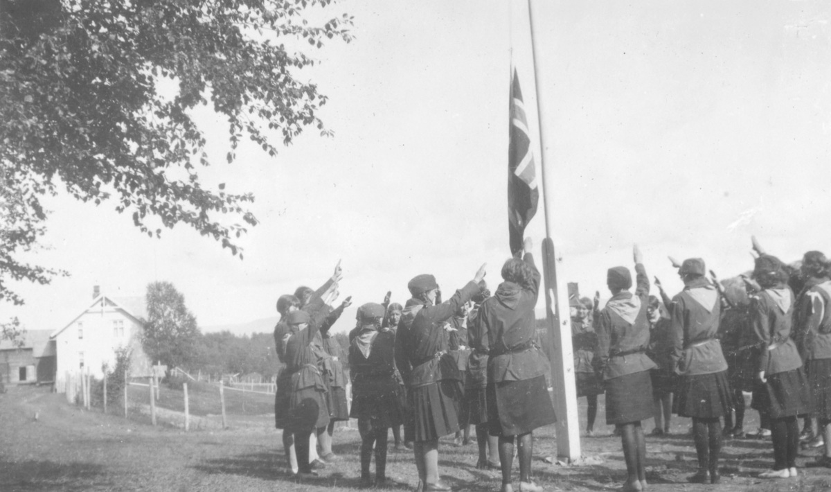 Speiderjenter på Kretsleir i Alta, 1928. Her blir det gjort flagghilsen en tidlig morgen. Jentene er kledt i uniformer. Til venstre er det et hvitt hus
