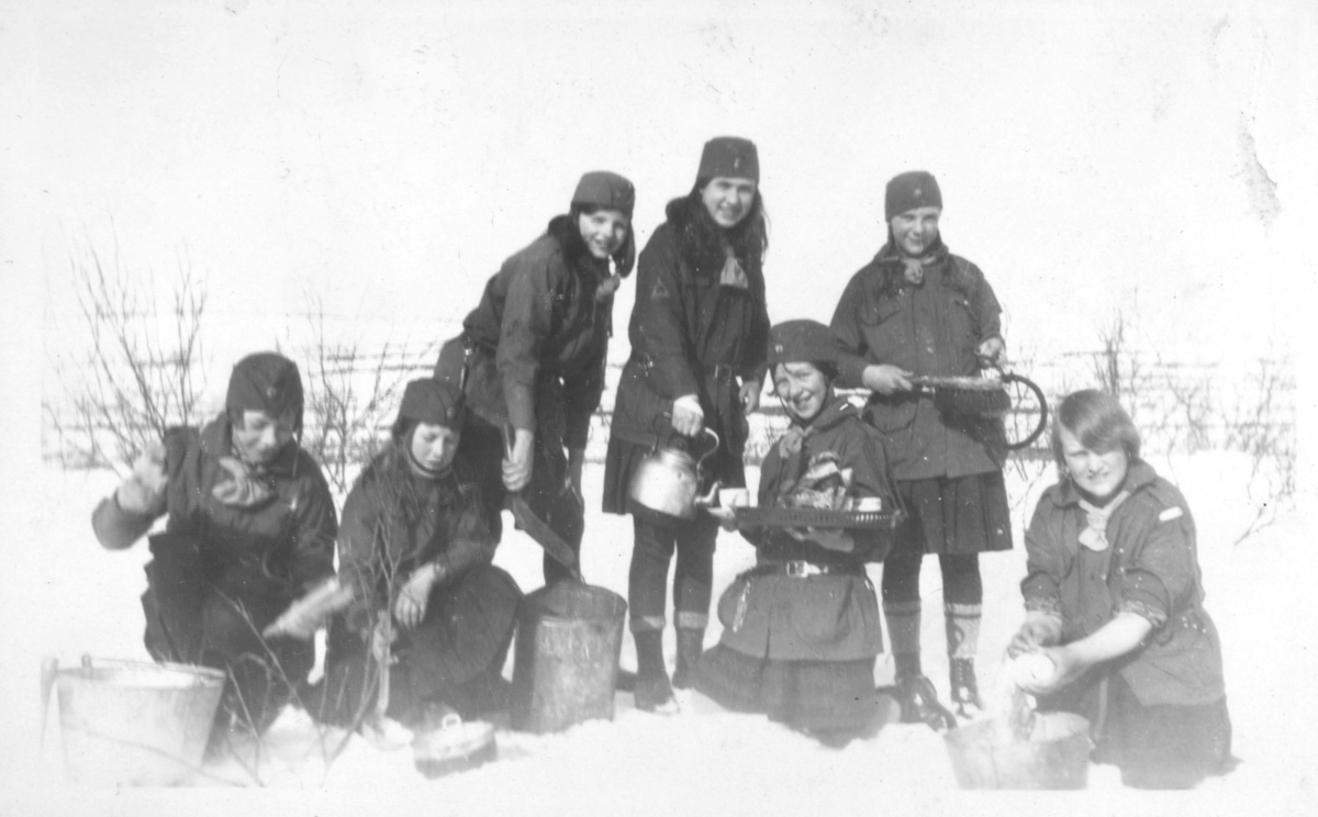 Speiderjenter på tur ved Hoels hytte i Tomaselv, vinteren 1930. På bildet er de ute i snøen. Fra venstre sitter: Aud Johannessen, Randi Feldt, Solveig Evanger, Arnhild Tøgersen, Johanna dahl, Astrid Pedersen, og Vera Hoel. Jentene er kledt i speideruniformer. De har bøtter, og kjeler, med seg ute i snøen. Arnhild skjenker fra en kaffekjele. Fjellet ses i bakgrunnen