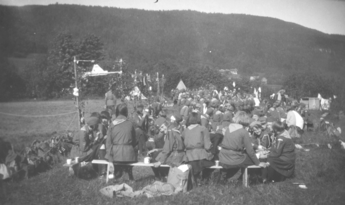 Speidere på pinseleir i Drammen, 1930. Speiderne er kledt i speideruniformer. De sitter på benker, hvor det også står kopper. Midt i bildet er det noen telt. Det er fjell i bakgrunnen av dem. Noen hus ligger til høyre i bildet