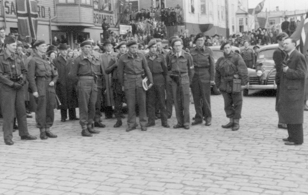 Frigjøringen i 1945. En gruppe soldater står oppstilt med store folkemasser bak seg oppover trapper og i dører og vinduer. En mann i sivil taler til soldatene.