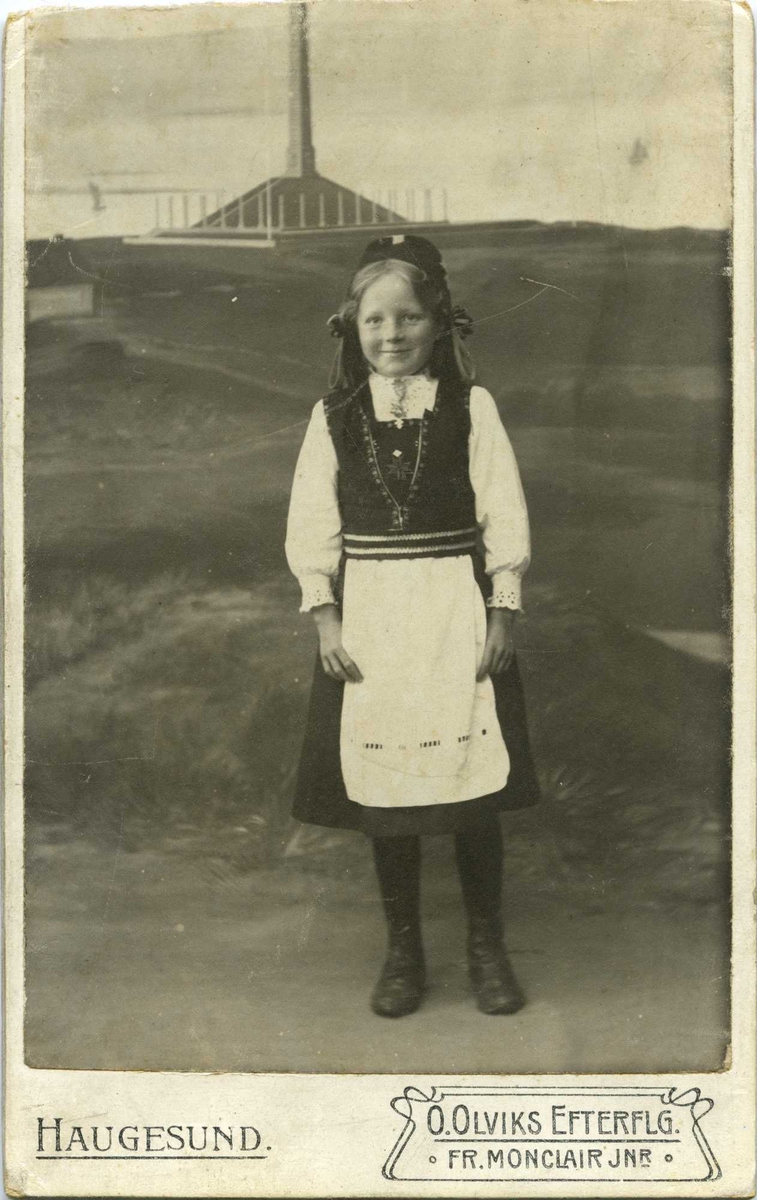 Portrett av jente i Hardanger-drakt. Jenta står foran en kulisse der Haraldstøtta er malt på.