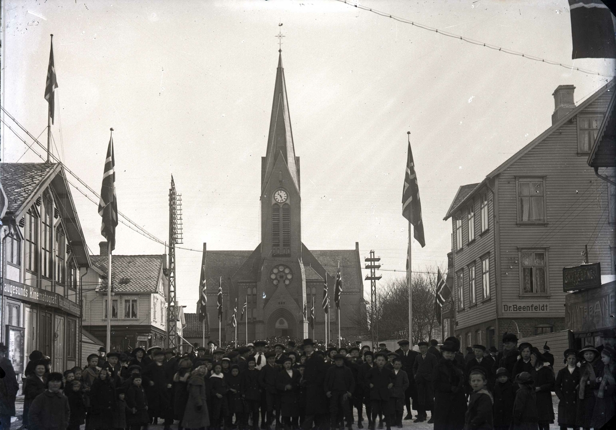 Stor folkemengde som kommer ut av dørene i  Vår Frelsers kirke, og vestover Torgbakken. Det flagges.